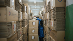 Factory warehouse aisle stacked with cartons of pickleball balls while a quality control inspector examines a sample ball before wholesale shipment